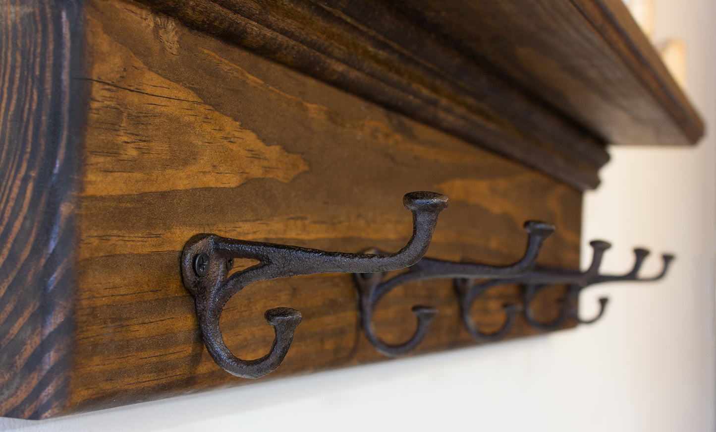 Close-up of a rustic wall-mounted coat rack with two cast iron hooks. The hooks are dark gray and mounted on a stained wooden board. The wood grain is visible, and the hooks are attached with screws.