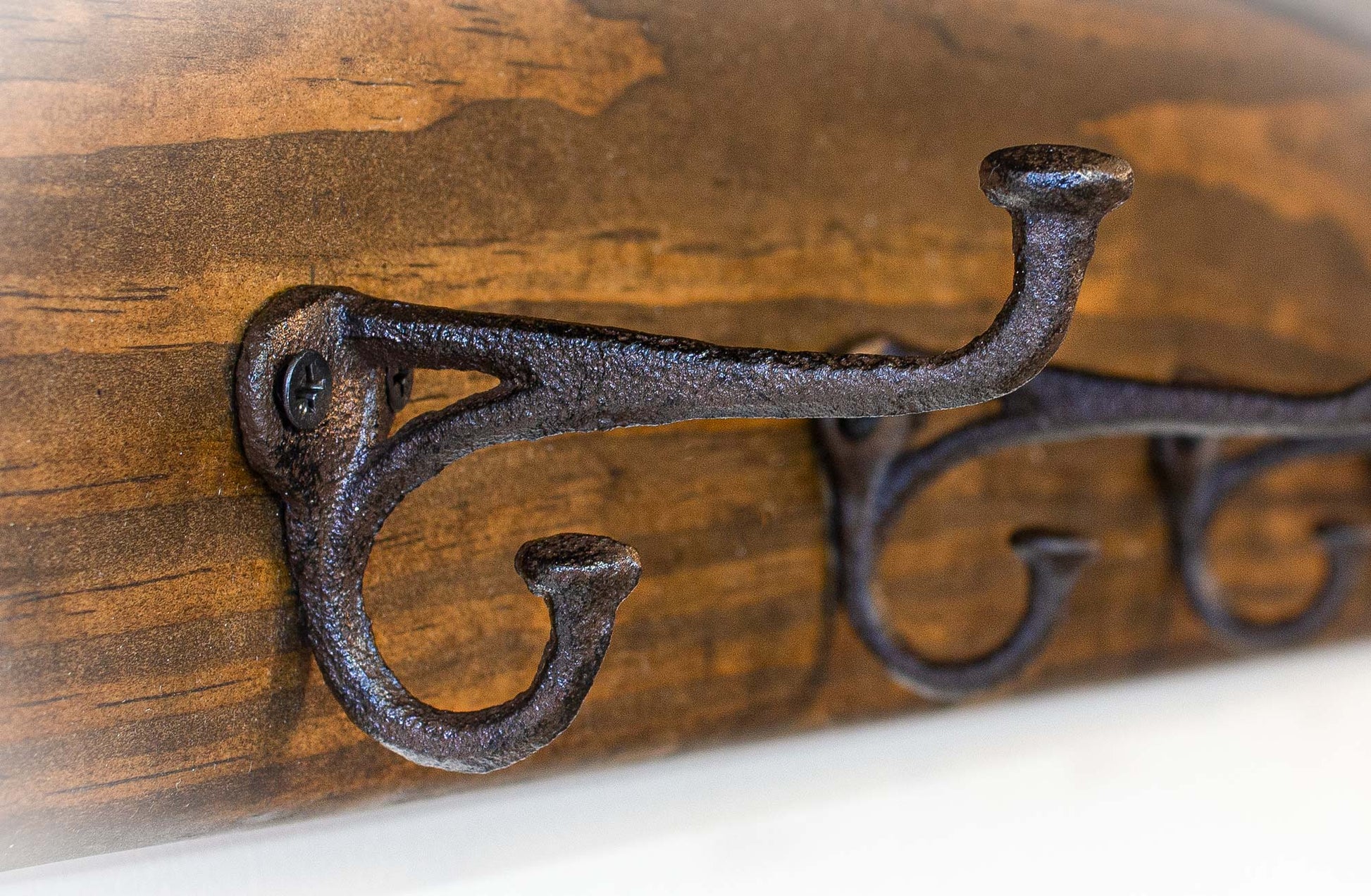 A rustic coat rack with three dark metal hooks mounted on a stained wooden board. The hooks have a curved design and are attached with screws. The wood grain is visible, adding to the natural aesthetic.