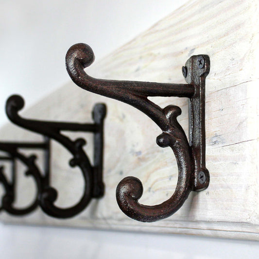 Close-up of a large decorative, dark brown metal coat hook with a curved design. The hook is mounted on a distressed, white-painted wooden board. The hook has a scroll-like design and is attached with screws.