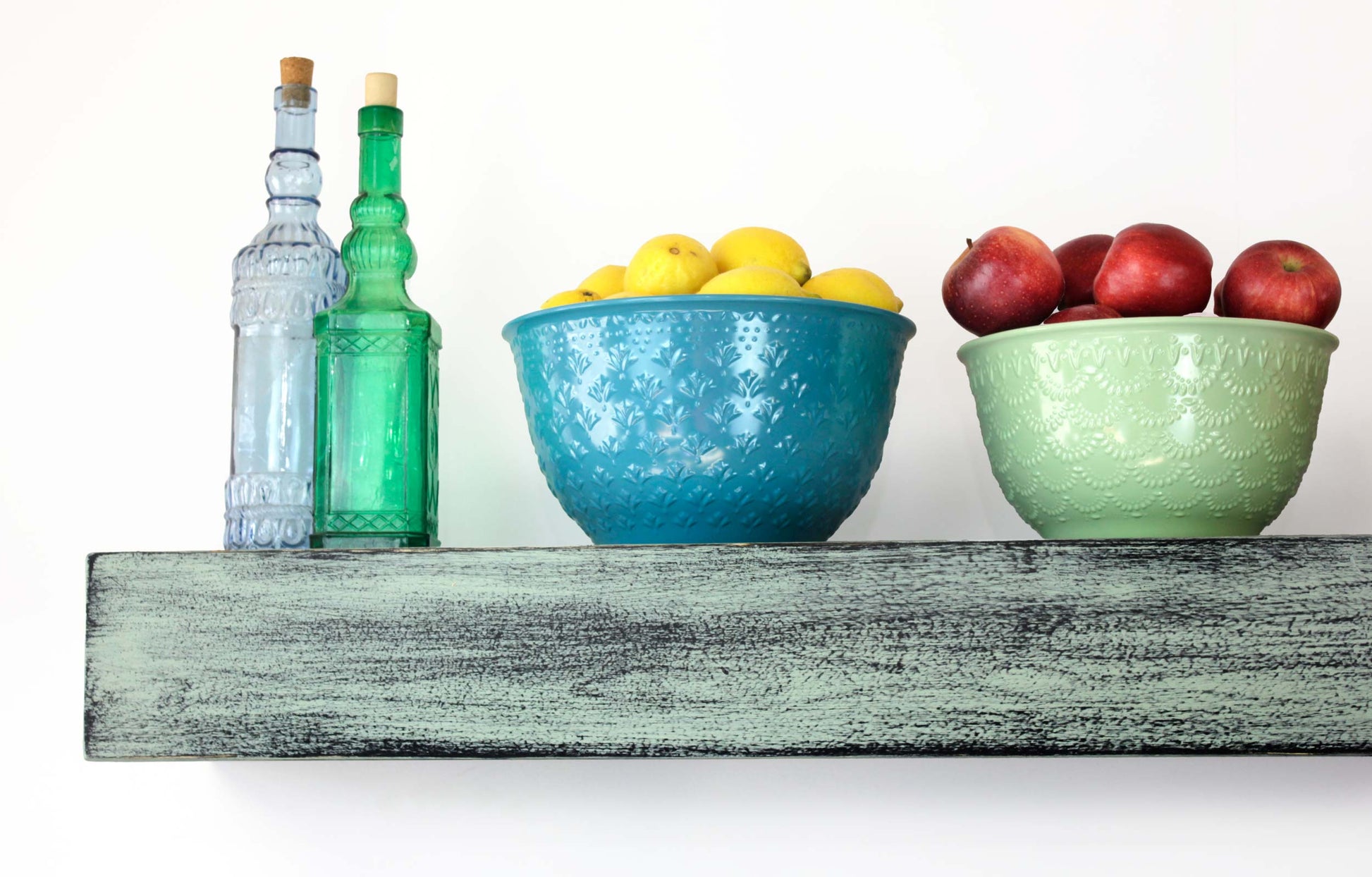 closeup detail of hand distressed paint sage green over black on a timber beam shelf with a bowl of yellow lemons, a bowl of red apples and 2 glass bottles sitting on the top