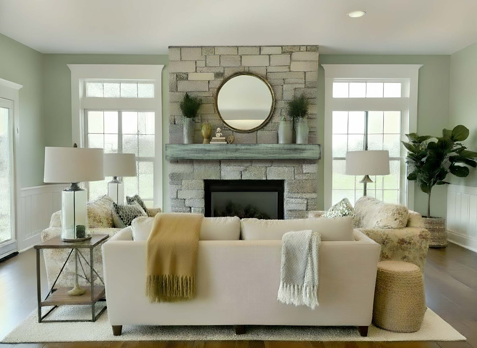 May include: A living room with a stone fireplace, a round mirror, and a light-colored sofa. Two lamps flank the sofa, and a side table is on the left. A tan throw blanket and a gray throw blanket are draped over the sofa.