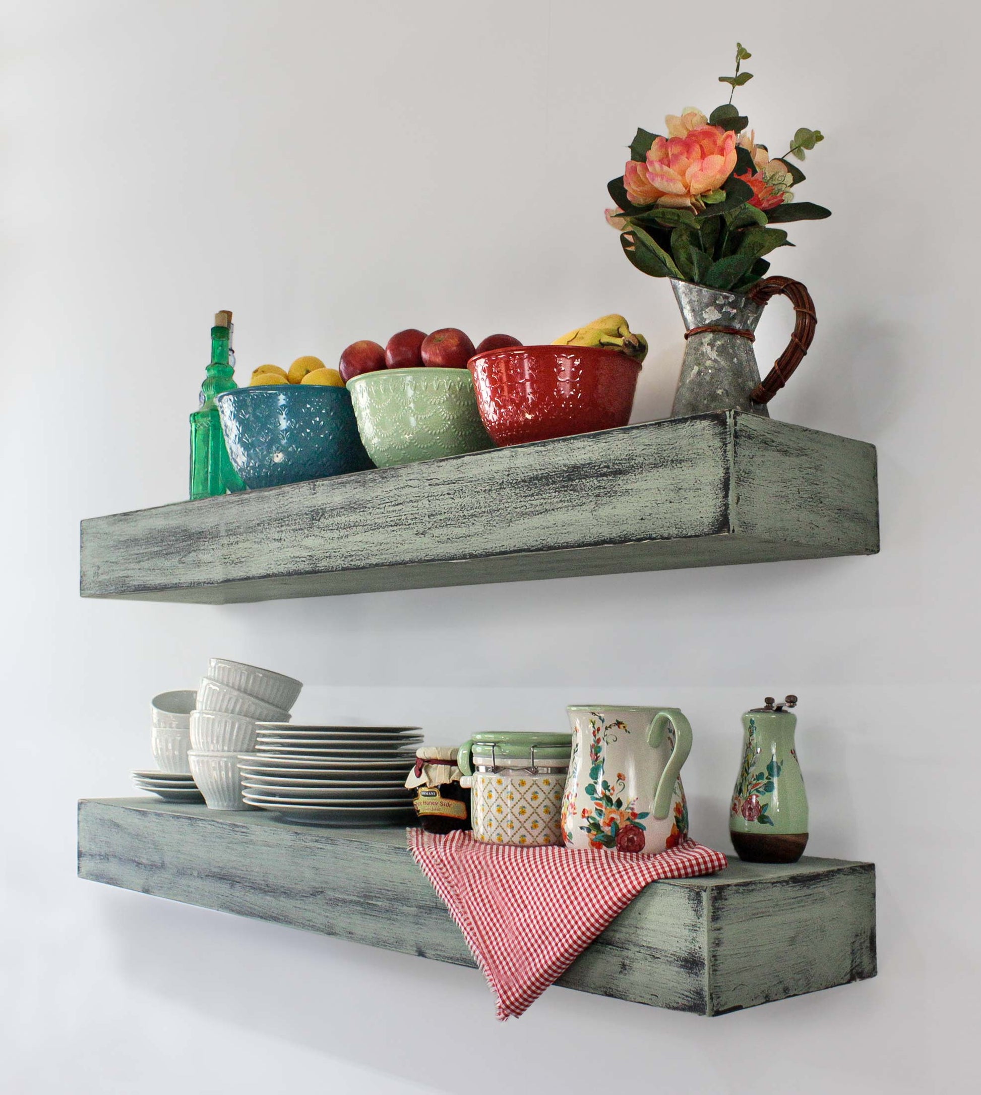 Two rustic, distressed, turquoise shelves display kitchenware. The top shelf holds a green bowl of apples and a red bowl. The bottom shelf has a jar of preserves, a patterned jar, a floral pitcher, and a stack of white plates. Perfect look for a retro 1940s and 1950s kitchen decor.