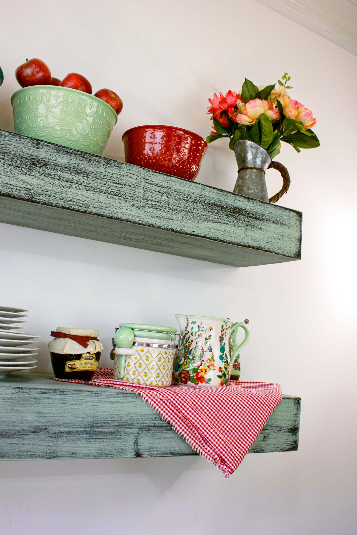 A rustic farmhouse 1940s - 1950s wooden shelf displays a collection of colorful bowls filled with fruit and a decorative vase with flowers. The bowls are turquoise, green, and red. Two glass bottles are on the left.
