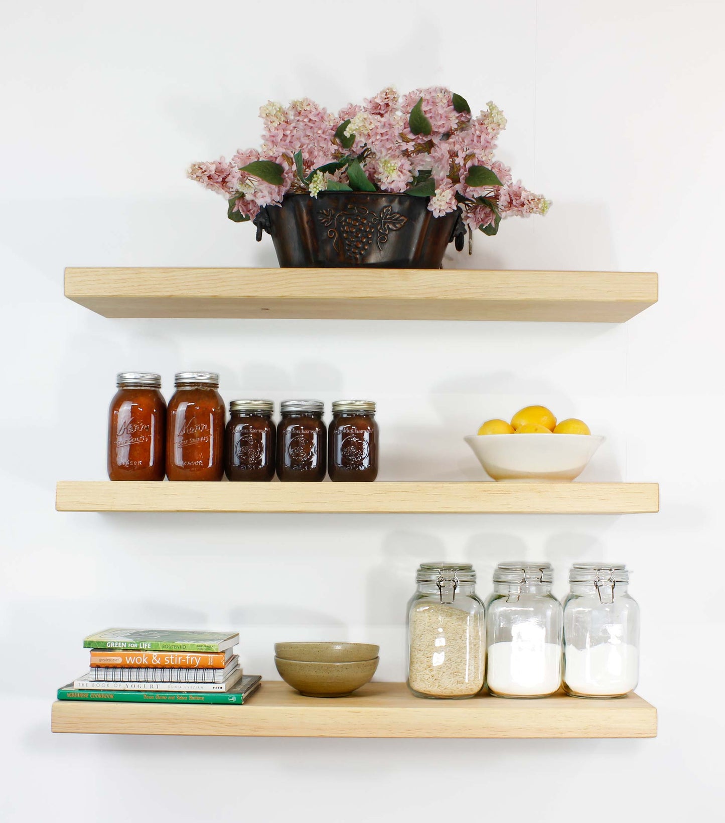Three wooden shelves against a white wall display various kitchen items. The top shelf holds a decorative metal planter with pink flowers. The middle shelf has jars of preserves and a bowl of lemons. The bottom shelf has cookbooks, a bowl, and jars of dry goods.
