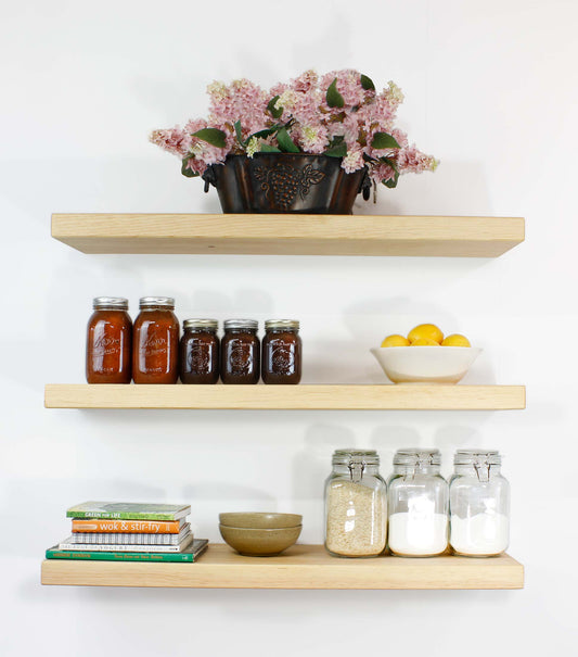 Three wooden shelves against a white wall display various kitchen items. The top shelf holds a decorative metal planter with pink flowers. The middle shelf has jars of preserves and a bowl of lemons. The bottom shelf has cookbooks, a bowl, and jars of dry goods.