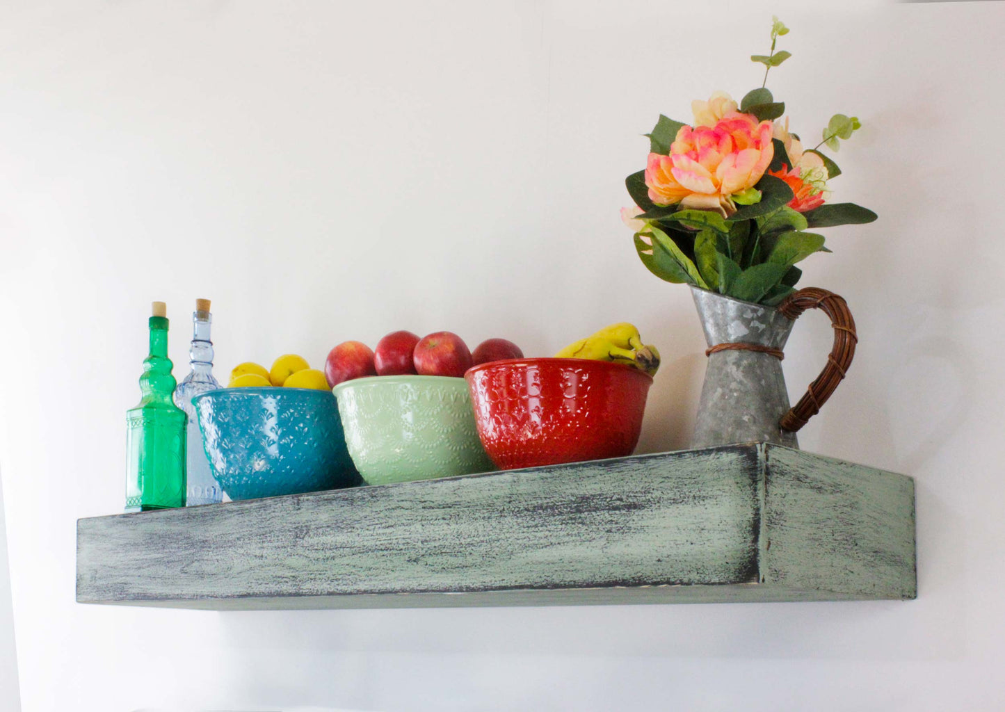 A rustic wooden shelf displays decorative glass bottles and bowls filled with fruit. The bottles are blue and green with cork stoppers. A turquoise bowl holds lemons, and a green bowl contains red apples.