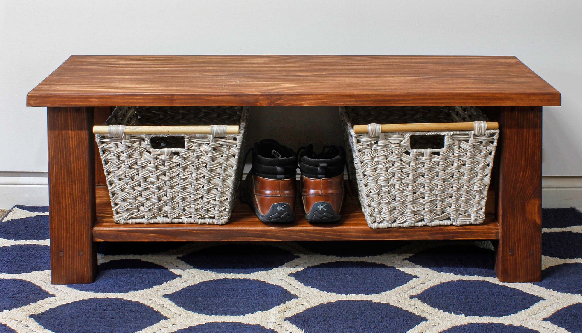 May include: A rectangular wooden coffee table with a dark brown finish. It features a lower shelf with two woven baskets and a pair of brown leather shoes. The table sits on a blue and white patterned rug. The rug, shoes, and baskets are not included in the purchase. Shows how well the bench can store items attractively underneath out of the way.